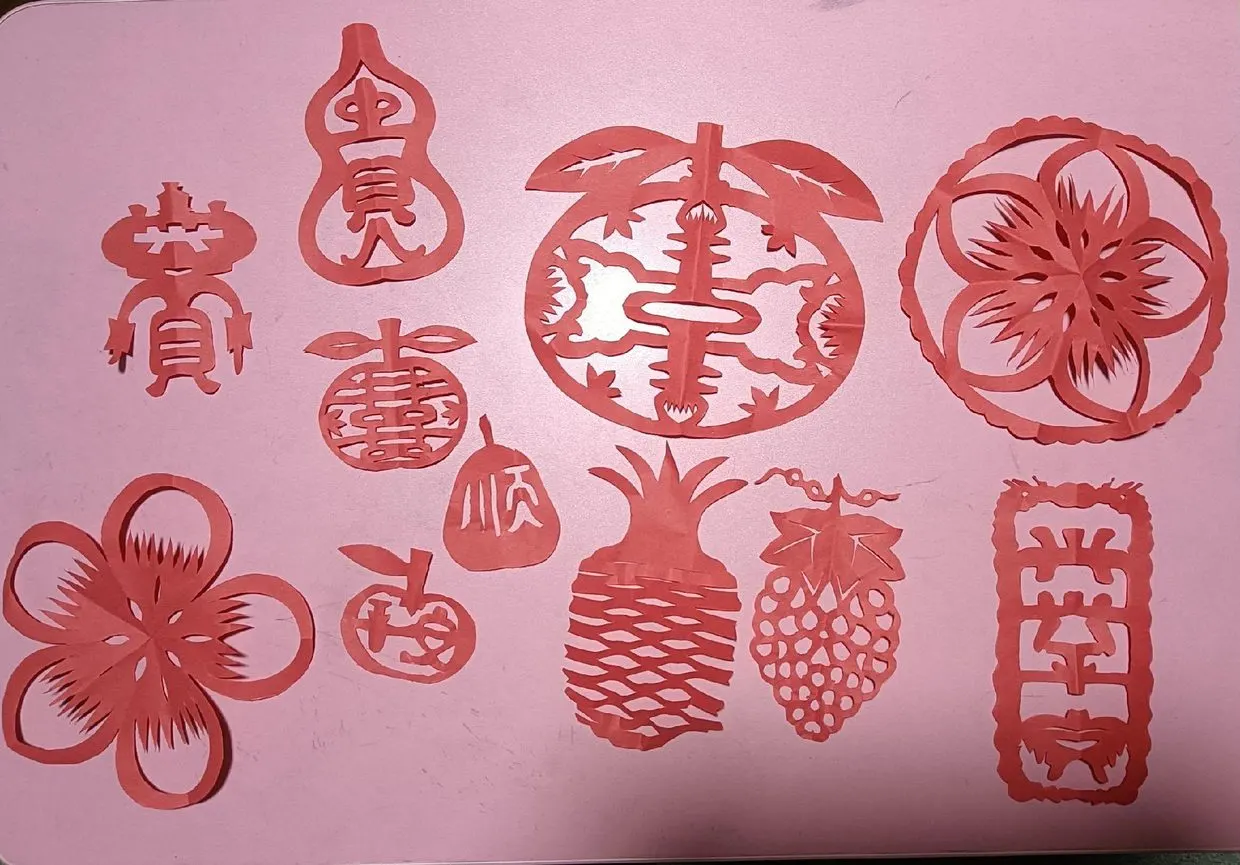 一组红纸剪纸:葫芦、桃、菠萝、葡萄与「平安」字样,平铺成一版。图源:抖音 @潮州剪纸黄朝阳(跃然斋文化传播),2023 年 9 月 29 日。