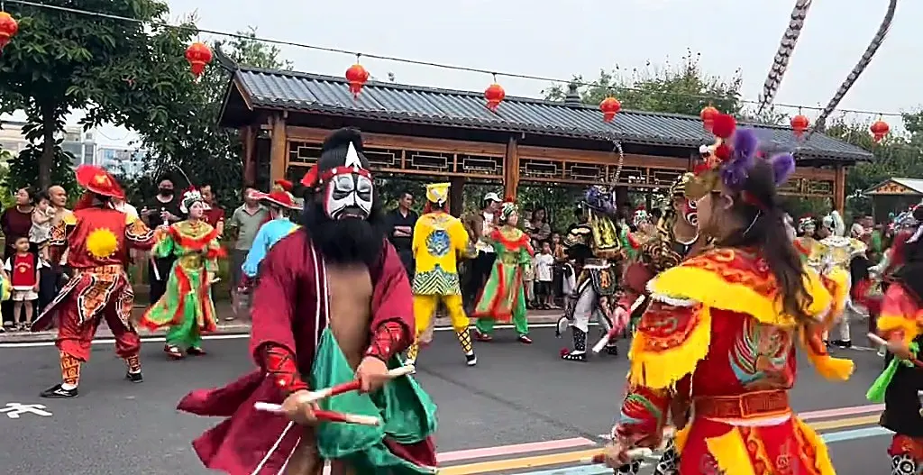一名黑脸长须、红袍的队员双手各执一根木槌站在街心,身后是彩绘长廊与红灯笼