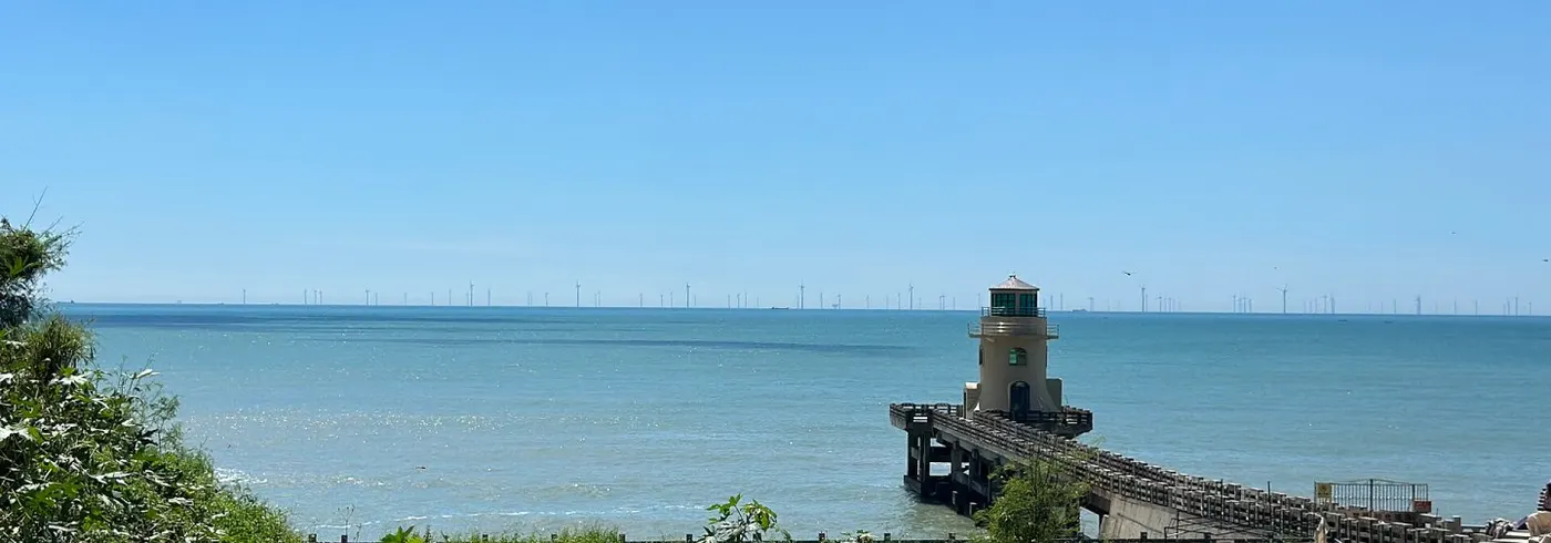 海边小灯塔立在石砌防波堤尽头,远处海平线排着一列海上风机
