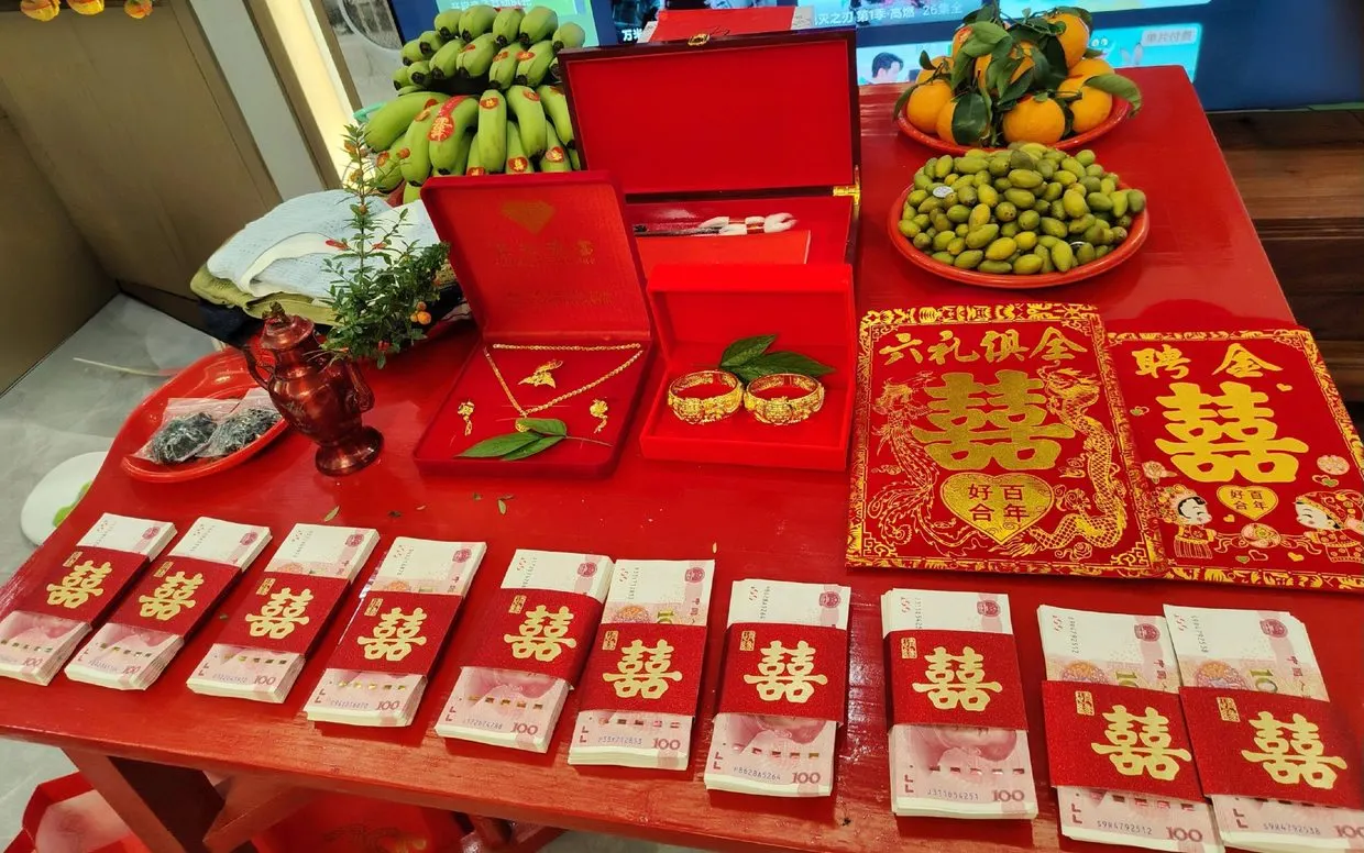 定亲的聘礼桌：龙凤镯、香蕉、柑橘、青橄榄、成叠红包，右边压着「六礼俱全」红帖。图源：抖音 @林～青娘（喜娘），7 月 3 日。