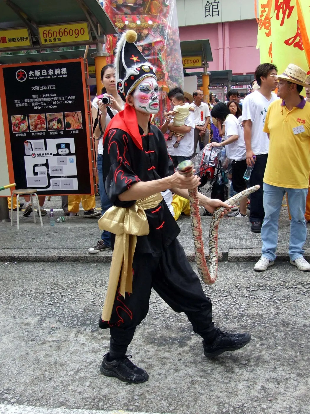 一人穿黑衣,手里垂着一条浅色布蛇,站在街边台阶上