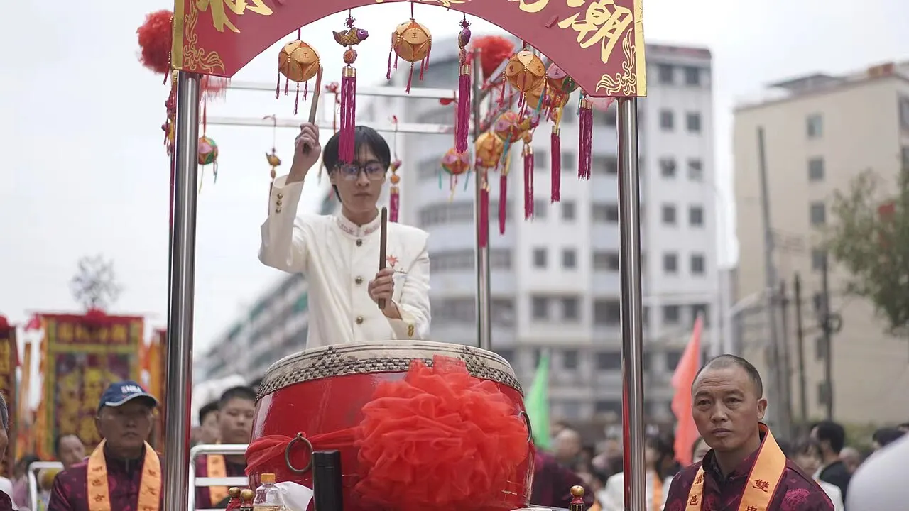营老爷队伍中,一人立于鼓亭击鼓,两侧人员佩红绶带